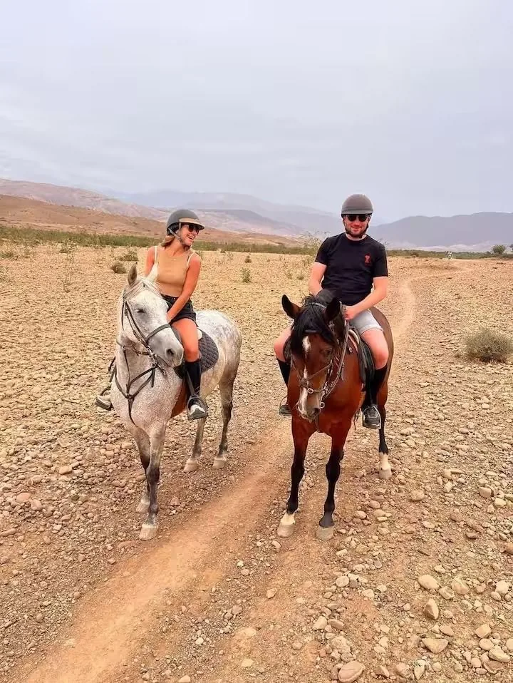 Two riders in helmets on grey and bay horses on a stony desert track with dry hills behind